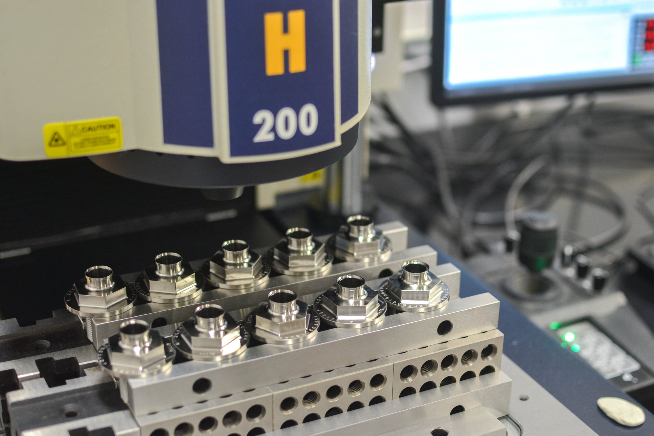 Close-up of metal parts in a precision engineering context, positioned under an industrial machine marked "h 200" in an inspection lab.