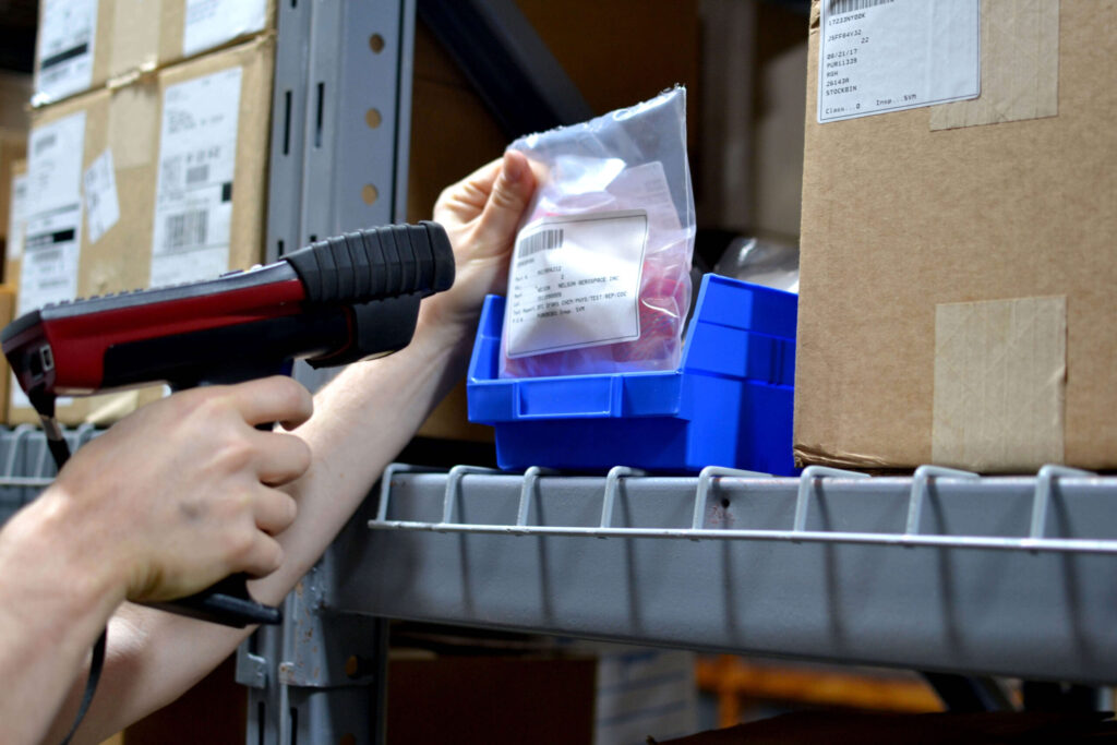 A worker scans a barcode on a packaged item in a warehouse using a handheld tooling scanner.