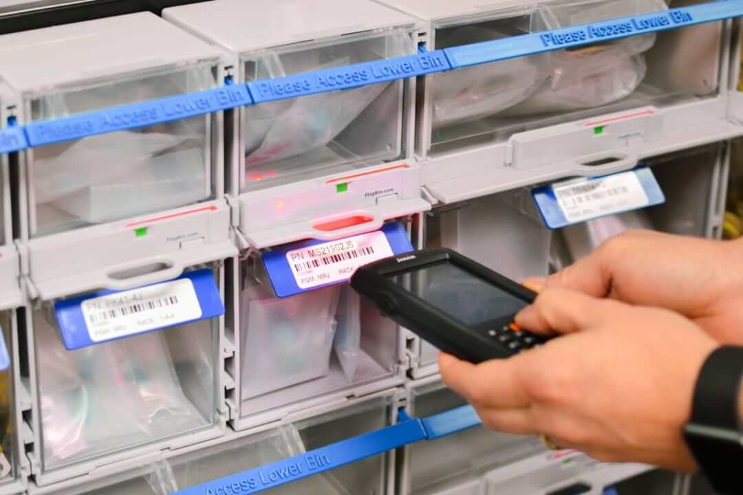 A person uses a handheld scanner on barcodes of storage bins containing fluid fittings in a well-organized inventory system.