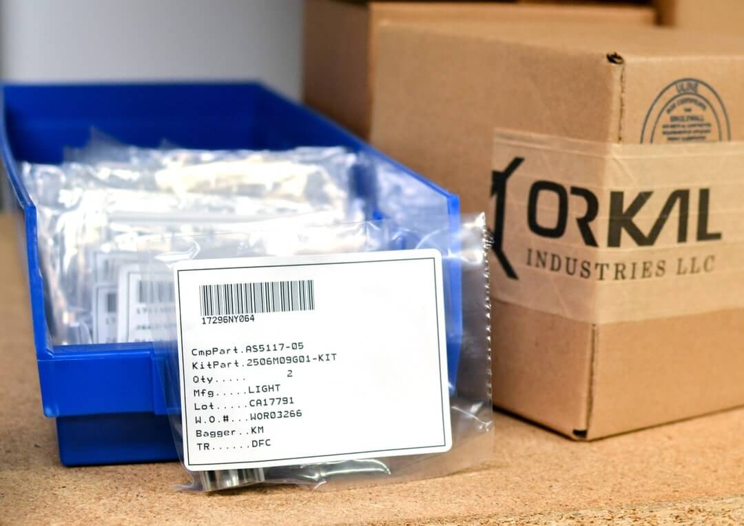 A clear plastic bag with a labeled aerospace parts kit sits in a blue bin beside a cardboard box marked "Forkal Industries LLC.