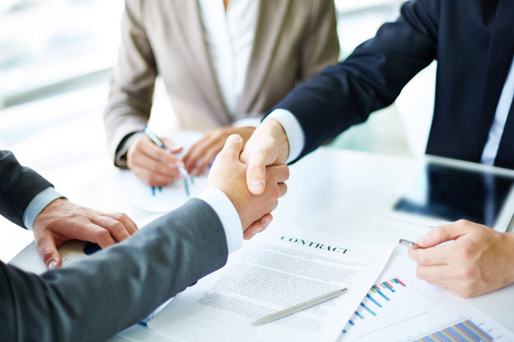 Two individuals shaking hands over a desk with a contract, in the presence of others, indicating a business agreement.