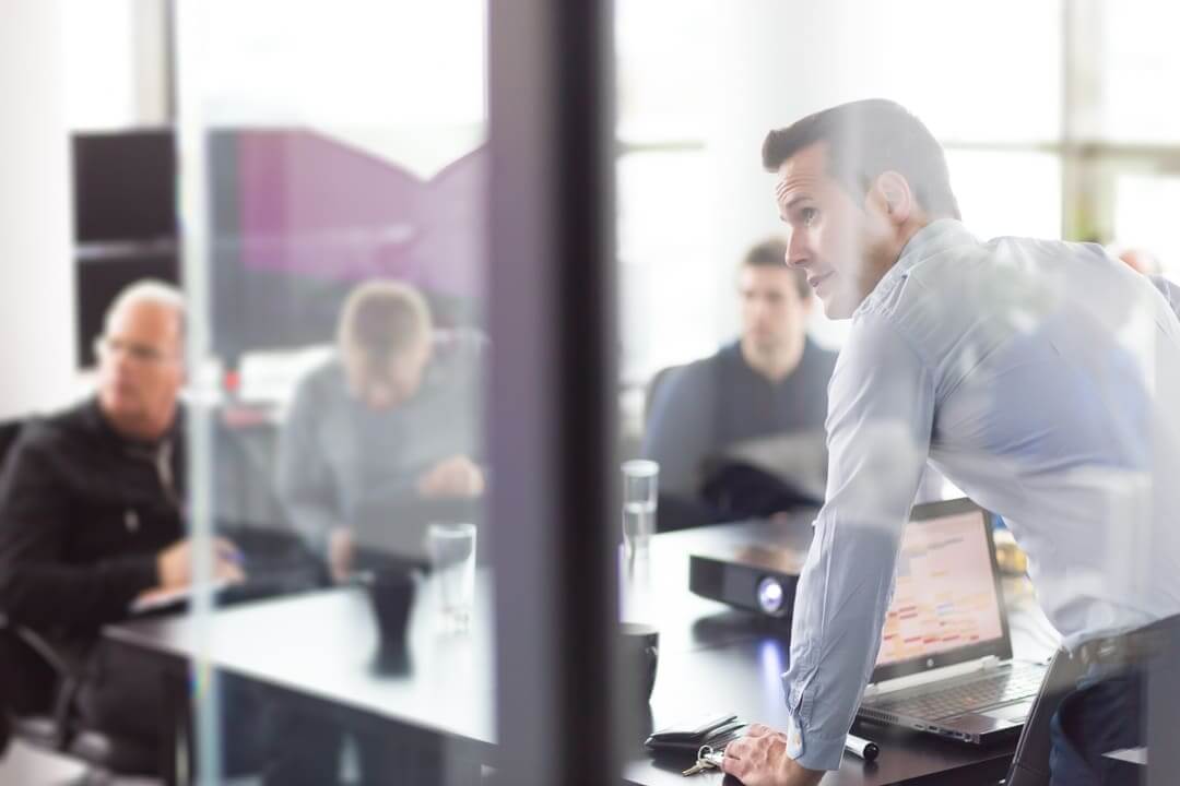 A man in a business meeting, standing and leaning on a table with blurred colleagues in the background.