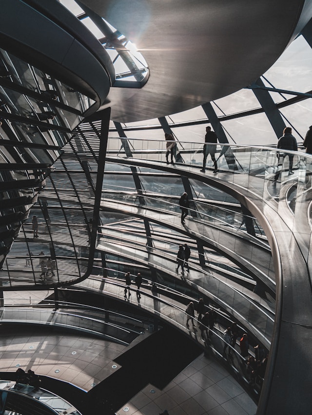 Interior of a modern building with curved glass panels and metal beams, featuring multiple levels with people walking on spiral walkways, designed under strict quality assurance protocols.